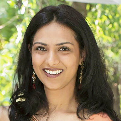 The image shows a woman with dark hair smiling at the camera. She has makeup on her face and is wearing a sleeveless top with a necklace.