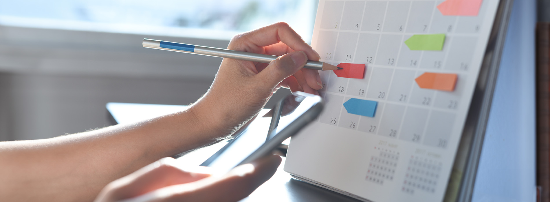 The image shows a person writing on a paper calendar with a pen, surrounded by other papers and a laptop keyboard, suggesting an organized workspace.