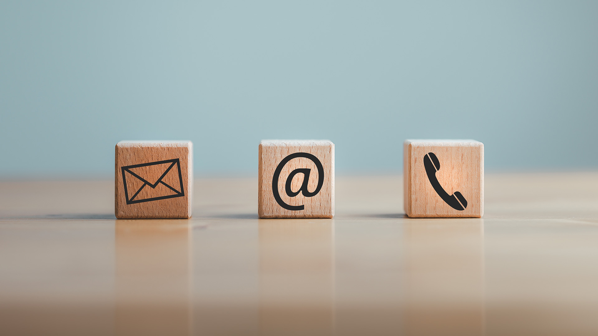 The image shows three wooden blocks with icons representing email, phone call, and mail, arranged diagonally on a surface against a blurred background.