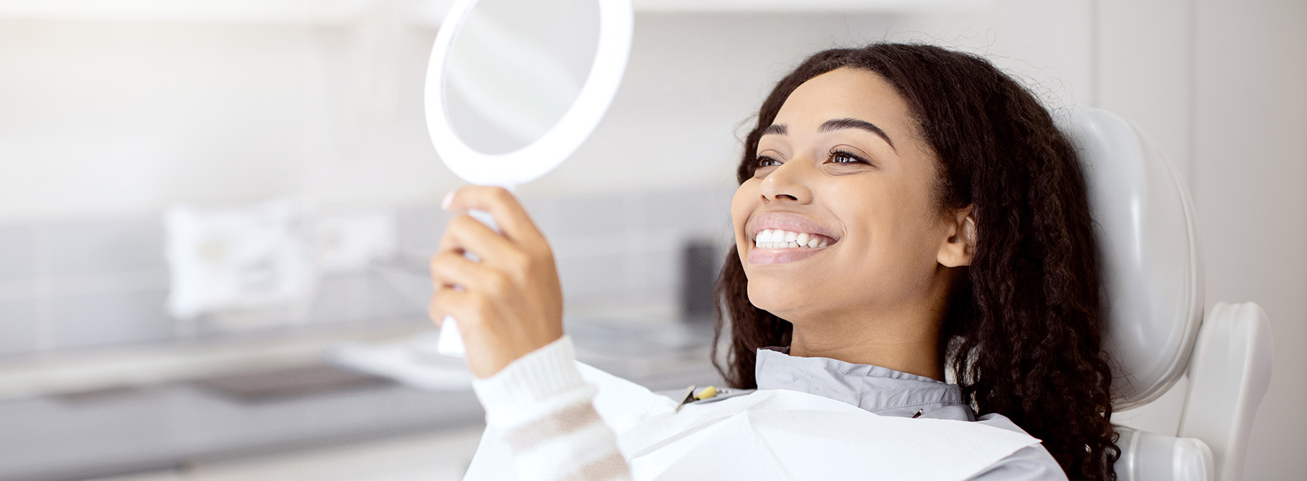 A woman smiling while holding a mirror in front of her face, with a dental chair visible in the background.