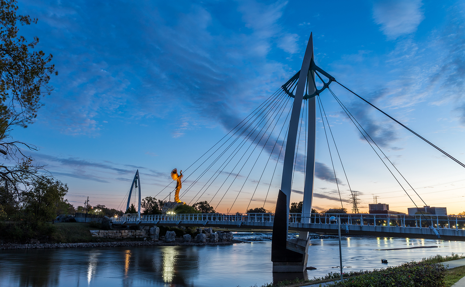 An evening view of a pedestrian bridge over a river at dusk with a city skyline in the background.