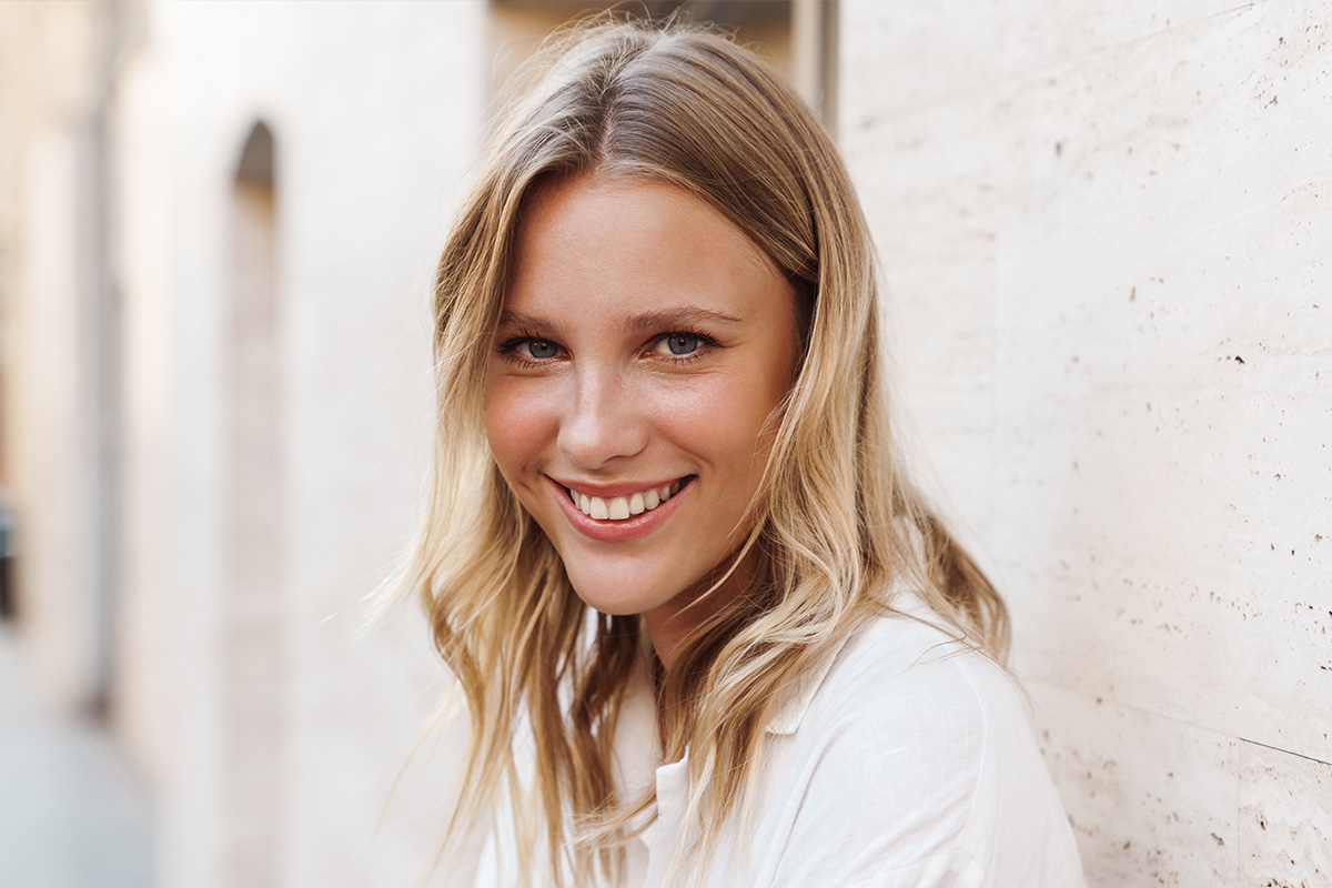 The image features a woman with blonde hair, wearing a white top, smiling at the camera while posing against a brick wall.