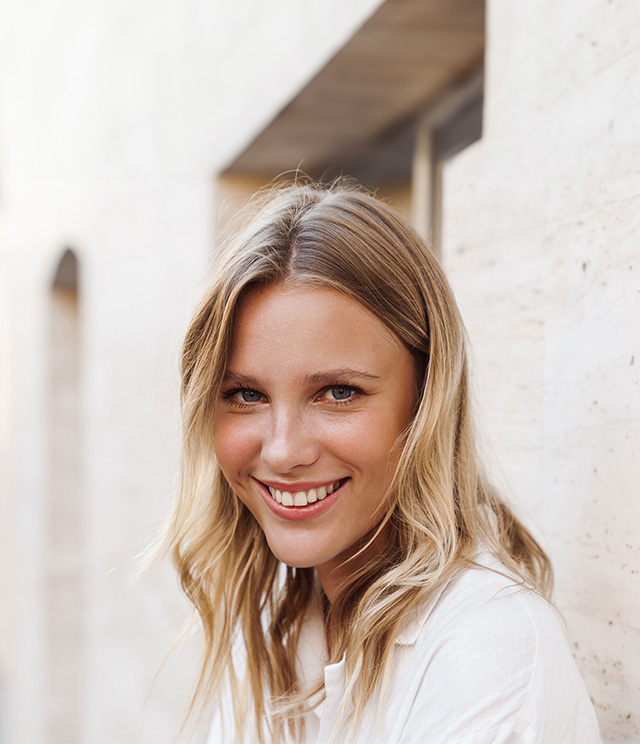 A young woman with blonde hair smiles at the camera against a backdrop of a beige brick wall.