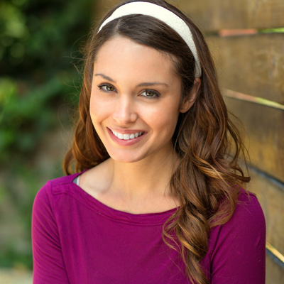 A woman with long brown hair, wearing a pink top and a white headband, smiling at the camera.