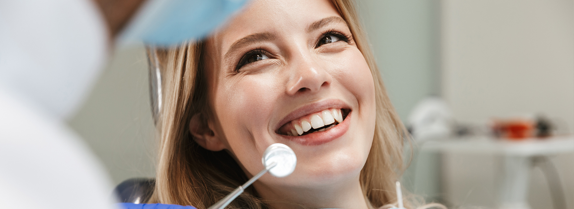 A smiling woman with a face mask seated at a dental chair, receiving dental treatment, with a dentist standing behind her holding a mirror, both in a dental office setting.