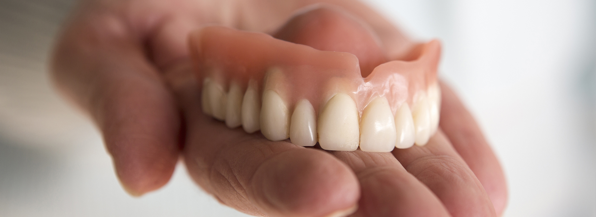 The image shows a person holding up a set of dentures with their hand, showcasing the upper teeth of the denture.