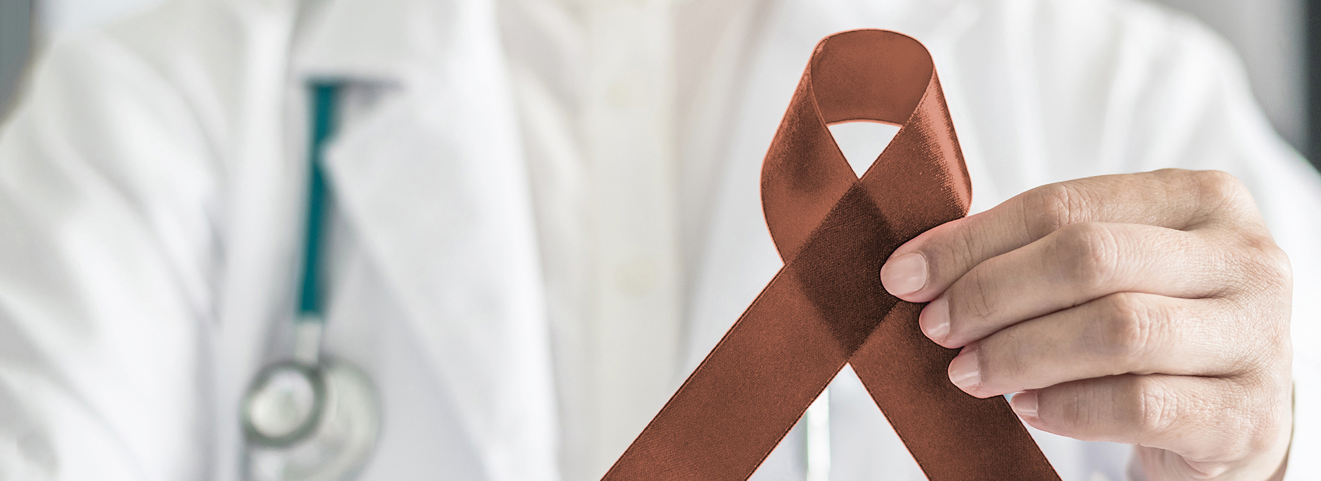 An adult male wearing a white lab coat holds up a red ribbon with a white cross, symbolizing awareness for a medical condition, against a grey background.