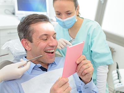 The image shows a man sitting in a dental chair with a pink card held up in front of him, while a female dental professional stands behind him, smiling and holding a mirror.