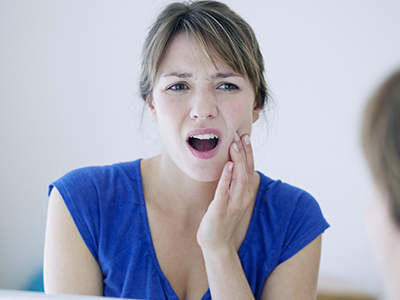 A woman with her mouth open, holding her hand to her face, appears to be reacting with surprise or concern while looking at a reflection in a mirror.