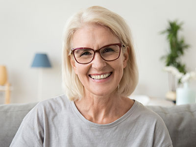 The image shows a woman with blonde hair and glasses smiling at the camera. She appears to be indoors, possibly in a living room setting.