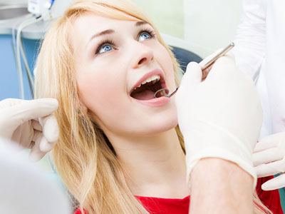 The image shows a woman sitting in a dental chair with her mouth open wide, receiving dental treatment, with a dental professional wearing gloves and a face mask performing the procedure.