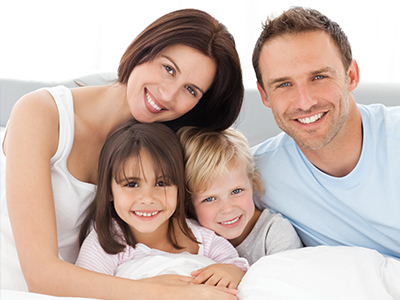 The image depicts a family of four adults and two children posing for a photograph on a bed with a white comforter, smiling at the camera.