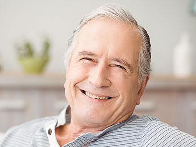 The image shows a smiling older man with gray hair, wearing a blue shirt, seated comfortably in a chair.