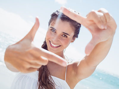 A woman with long hair is smiling while holding up her hand and fingers to create a frame around the camera lens.